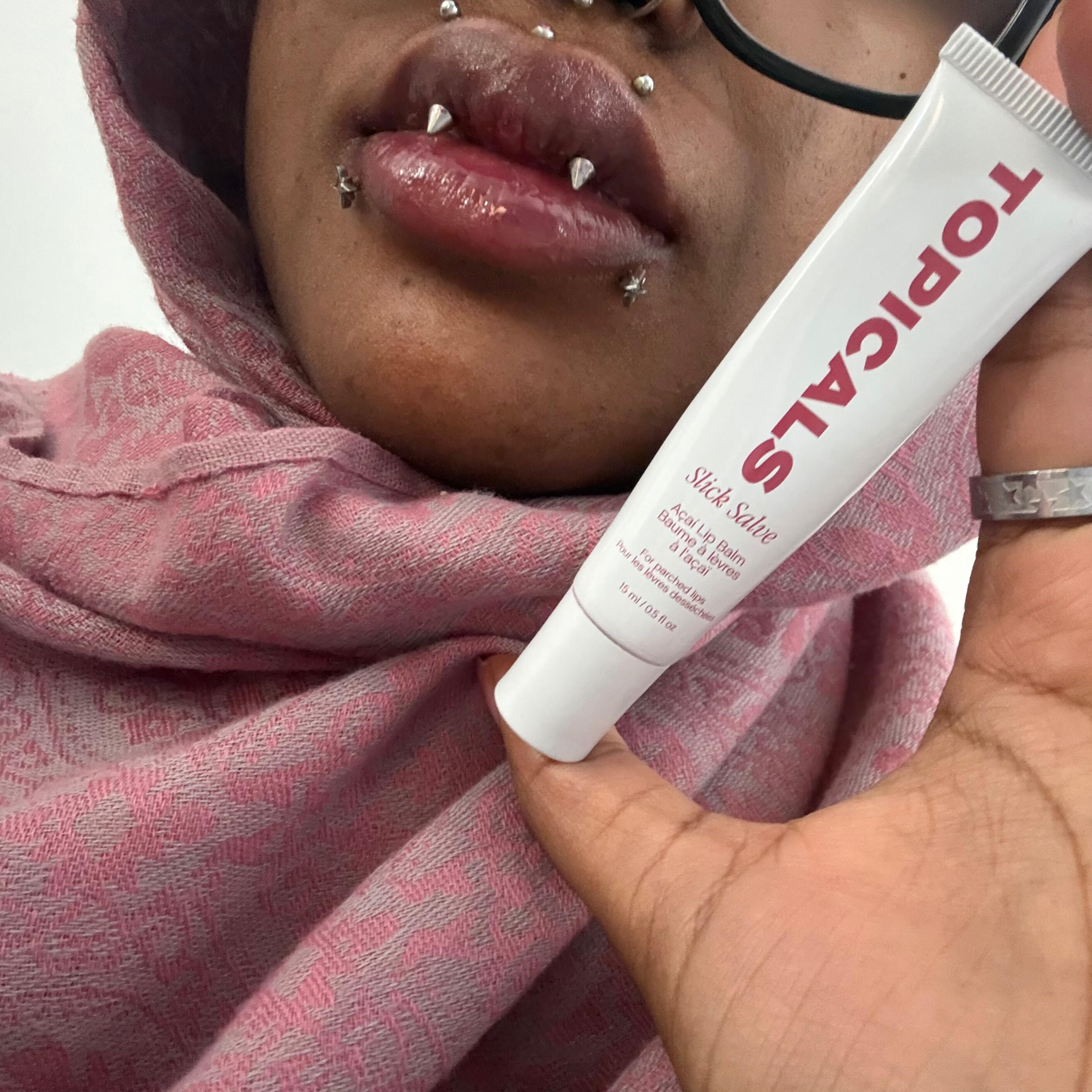 Me Holding Topicals Lip Salve Brown face cut off from the top down stopping at the lips, showing angel fang and medusa and snakebite piercings, holding a tube of the topicals lip salve in açaí and wearing a pink patterned pashmina scarf and a stainless steel ring with stars engraved on